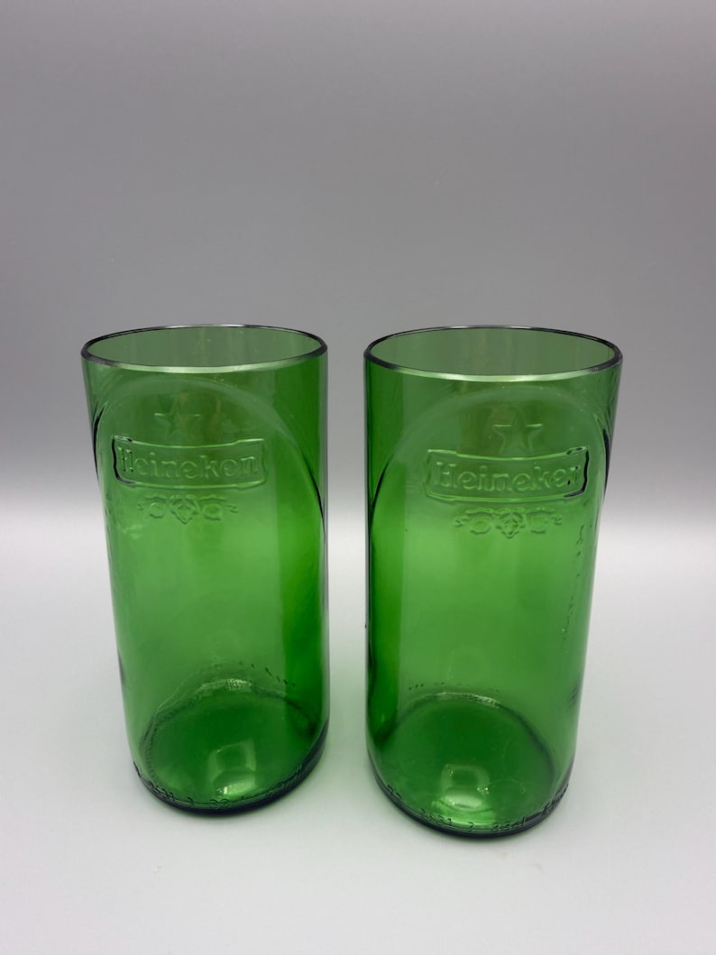 May include: Two cylindrical drinking glasses crafted from green glass bottles. Each glass features the embossed Heineken logo and a star symbol on the front surface. The glasses have a smooth, straight-sided design with a clear, open top rim.