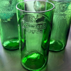 May include: Three cylindrical drinking glasses crafted from green glass bottles. Each glass features the embossed Heineken logo, a star symbol, and decorative leaf motifs on the front surface. The glass has a smooth, translucent finish.