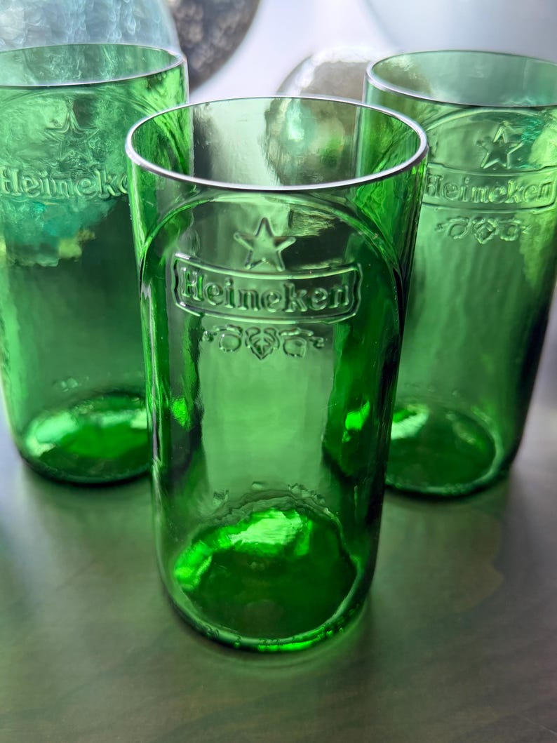 May include: Three cylindrical drinking glasses crafted from green glass bottles. Each glass features the embossed Heineken logo, a star symbol, and decorative leaf motifs on the front surface. The glass has a smooth, translucent finish.