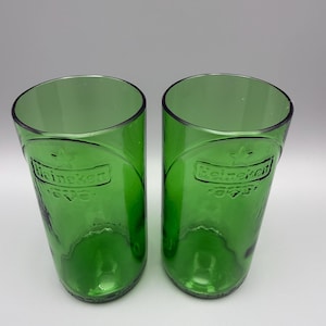 May include: Two cylindrical drinking glasses crafted from green glass bottles. Each glass features the embossed Heineken logo and a star symbol on the front surface. The glasses have a smooth, open top rim and a rounded base, maintaining the original green color and texture of the glass material.