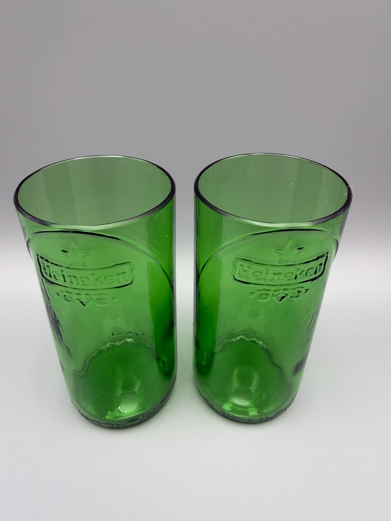 May include: Two cylindrical drinking glasses crafted from green glass bottles. Each glass features the embossed Heineken logo and a star symbol on the front surface. The glasses have a smooth, open top rim and a rounded base, maintaining the original green color and texture of the glass material.