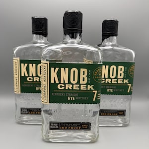 May include: Three empty glass bottles of Knob Creek Kentucky Straight Rye Whiskey. Each bottle features a dark green label with gold text, a black cap, and embossed lettering on the glass base. The labels state 100 Proof and 7 years aged.