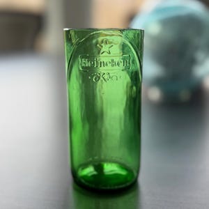 May include: A green glass tumbler crafted from a repurposed bottle, featuring the embossed Heineken logo and a star symbol near the rim. The cylindrical vessel has a smooth, translucent finish and a thick, rounded base, showing clear branding.