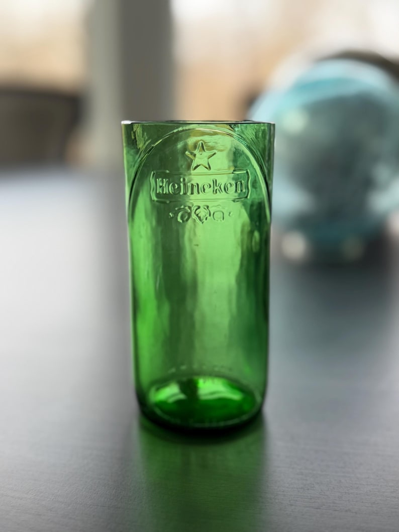 May include: A green glass tumbler crafted from a repurposed bottle, featuring the embossed Heineken logo and a star symbol near the rim. The cylindrical vessel has a smooth, translucent finish and a thick, rounded base, showing clear branding.