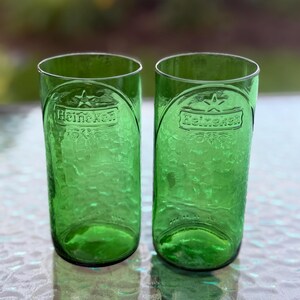 May include: Two cylindrical drinking glasses crafted from green glass, each featuring the embossed Heineken logo and a star symbol near the rim. The surface of the glasses has a textured, dimpled appearance, and the base is rounded.