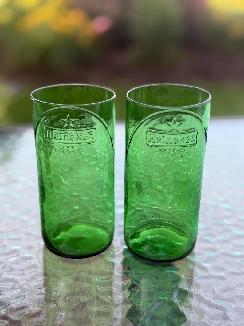 May include: Two cylindrical drinking glasses crafted from green glass, each featuring the embossed Heineken logo and a star symbol near the rim. The surface of the glasses has a textured, dimpled appearance, and the base is rounded.