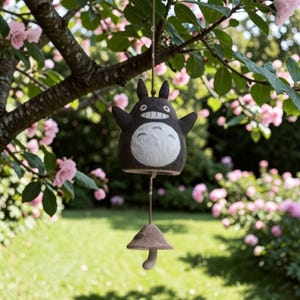 Puede incluir: Un carillón de viento Totoro en blanco y negro colgando de una rama de árbol. El carillón presenta una figura de Totoro sonriente con los brazos extendidos y un badajo en forma de hongo. El fondo incluye flores rosas y hierba verde.