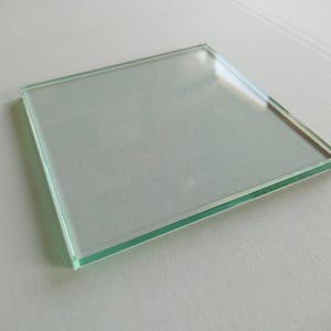 Peut inclure: Un dessous de verre carré en verre transparent avec des bords lisses et polis, présentant une légère teinte verte visible sur l'épaisseur du verre. La surface est plate et transparente, reflétant la lumière sur le dessus, avec un design simple.