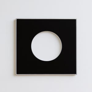 Könnte beinhalten: Ein quadratischer schwarzer Rahmen mit einer kreisförmigen weißen Öffnung in der Mitte. Der Rahmen hat einen dünnen silbernen Rand und ist vor einem weißen Hintergrund platziert. Das Design ist minimalistisch und modern.