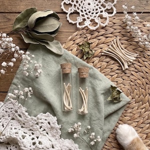 May include: Two glass vials with cork stoppers, each filled with thin, off-white sticks, rest on a sage green cloth. The image includes dried leaves, white lace, and a woven mat, creating a rustic, natural aesthetic. The vials are the focal point.