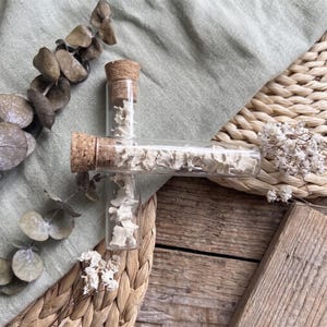 May include: Three small glass vials with cork stoppers, each filled with small, off-white bone fragments. The vials are arranged on a light green fabric, with dried eucalyptus leaves and white dried flowers. The background includes a woven basket and wooden surface.