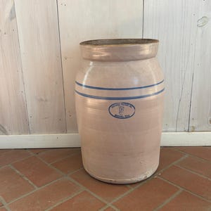 以下が含まれることがあります： 淡いピンク色の釉薬が施された、大きなアンティークの炻器の壺。2本の青いストライプと、数字の5が入った青い楕円形のエンブレムが特徴です。壺は円筒形で、わずかにフレアしたリムが付いています。