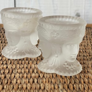Könnte beinhalten: Zwei Kerzenhalter aus mattiertem Glas. Jeder Halter hat ein detailliertes Gesichtsdesign mit dekorativem Kopfschmuck und einem ausgestellten Sockel. Die Kerzenhalter werden auf einer gewebten Oberfläche präsentiert, die ihre komplizierten Details und ihre elegante Form hervorhebt.