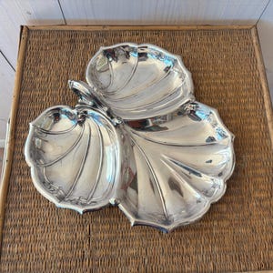 May include: A silver-colored, three-section serving dish in a leaf design. The dish has a polished, reflective surface and is placed on a woven, brown surface. The leaf-shaped sections are connected by a stem.