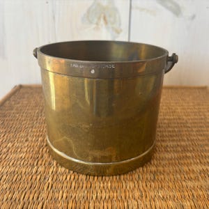 Puede incluir: Un cubo cilíndrico de latón con dos asas pequeñas y el texto "FAIRBANKS-MORSE" grabado en el borde. El cubo tiene un aspecto brillante y envejecido y está sobre una superficie tejida.