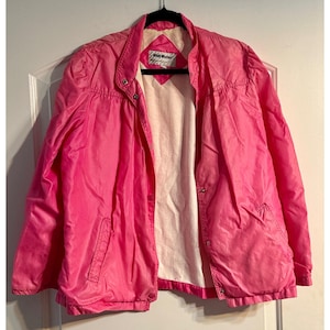 May include: A bright pink vintage jacket with a snap closure and a white interior lining. The jacket has a collar and two front pockets. The label reads "Windy Weather". The jacket is hanging on a black hanger.