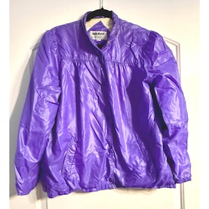 May include: A vibrant purple jacket with a stand-up collar and button closure. The jacket has a shiny, nylon-like material and features two front pockets. The jacket is hanging on a black hanger against a white door.