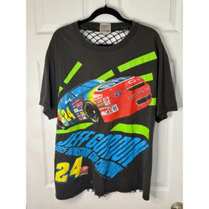Puede incluir: Camiseta negra con un gráfico de coche de carreras, el número 24 y el texto "JEFF GORDON 1995 WINSTON CUP CHAMPION". La camiseta tiene un aspecto desgastado y un cuello de red. El diseño presenta acentos azules, rojos y verdes.