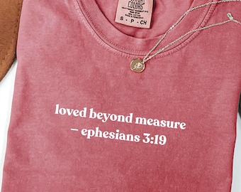 May include: A dusty rose Comfort Colors t-shirt with the text "loved beyond measure - ephesians 3:19" in white. A silver necklace with a small pendant rests on the shirt. The shirt is folded on a white surface.
