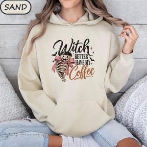 Peut inclure: Un sweat à capuche couleur sable avec une impression graphique représentant une main de squelette tenant une tasse de café avec un visage de citrouille. Le texte indique Witch BETTER HAVE MY Coffee, avec des étoiles et des chauves-souris.