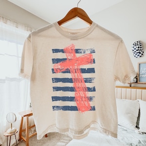 May include: A cream-colored short-sleeve t-shirt featuring a graphic print of a coral-colored cross layered over horizontal navy blue distressed stripes. The shirt is displayed on a wooden hanger against a neutral background.