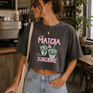 May include: A charcoal gray cropped t-shirt featuring a graphic print of two iced matcha drinks in green with pink text that reads Iced Matcha & Sunshine and all day every day. The shirt has short sleeves and a crew neckline, paired with light blue denim jeans.