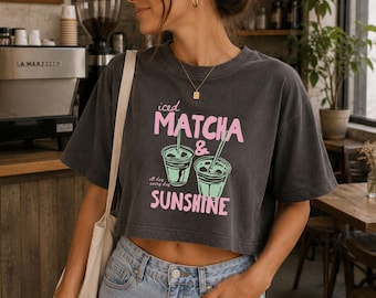 May include: A charcoal gray cropped t-shirt featuring a graphic print of two iced matcha drinks in green with pink text that reads Iced Matcha & Sunshine and all day every day. The shirt has short sleeves and a crew neckline, paired with light blue denim jeans.