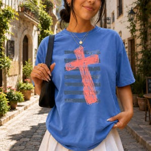 May include: A blue short-sleeve t-shirt featuring a large, textured coral-colored cross graphic centered over horizontal dark gray stripes. The shirt has a crew neckline and a relaxed fit, showcasing a bold, distressed-style print on the front.
