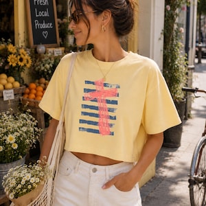 May include: A pale yellow short-sleeve crop top featuring a graphic design of a bright pink cross layered over horizontal navy blue distressed stripes. The garment has a crew neckline and a boxy fit, constructed from a solid-colored cotton fabric.