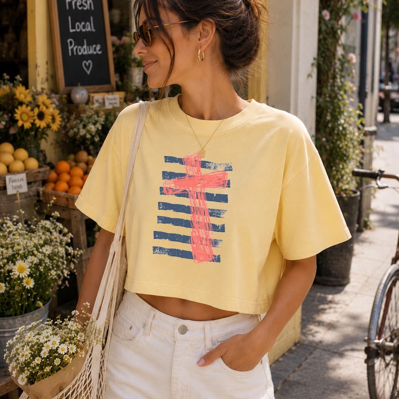 May include: A pale yellow short-sleeve crop top featuring a graphic design of a bright pink cross layered over horizontal navy blue distressed stripes. The garment has a crew neckline and a boxy fit, constructed from a solid-colored cotton fabric.