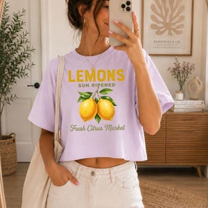 Op de afbeelding: Een lichtpaars T-shirt met korte mouwen en een afbeelding van twee gele citroenen met groene bladeren. De tekst luidt LEMONS SUN RIPENED en Fresh Citrus Market in gele en groene letters. Het T-shirt heeft een ronde hals en een wijde pasvorm.