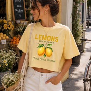 Op de afbeelding: Een lichtgeel kort T-shirt met een grafische print van twee citroenen met groene bladeren. De tekst luidt LEMONS SUN RIPENED en Fresh Citrus Market in een mix van vette en cursieve lettertypes. Het T-shirt heeft korte mouwen en een ronde hals.