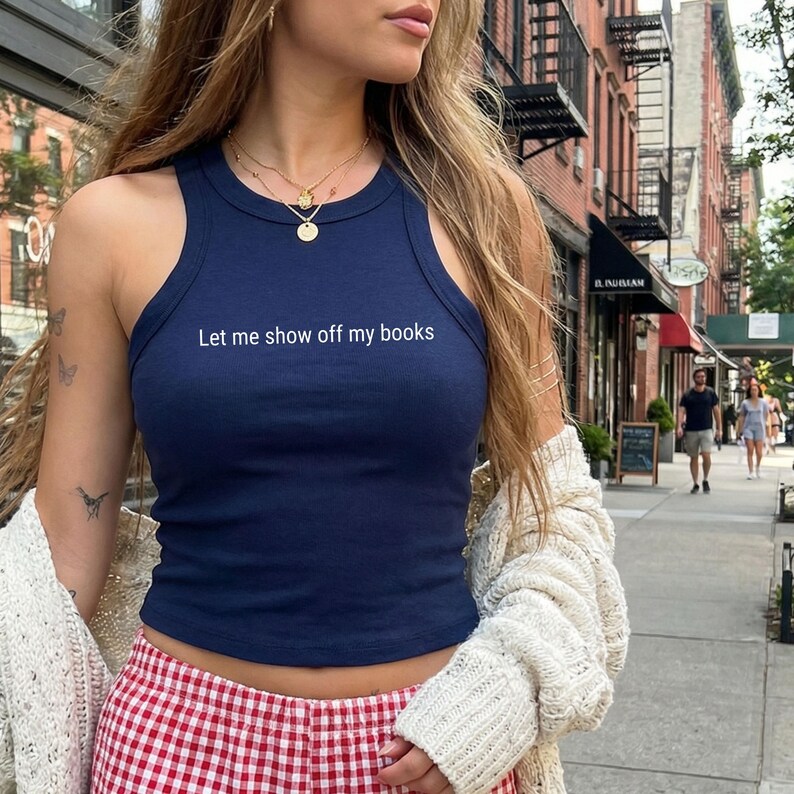 May include: A dark blue ribbed racerback tank top featuring the white text Let me show off my books printed on the chest. The garment is worn with a cream-coloured knit cardigan and red and white chequered bottoms, accessorised with layered gold necklaces.