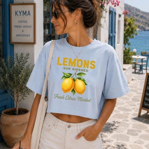 Op de afbeelding: Een lichtblauw, kort T-shirt met korte mouwen en een afbeelding van twee gele citroenen met groene bladeren. De tekst luidt LEMONS SUN RIPENED en Fresh Citrus Market in gele en groene letters. Het shirt heeft een ronde halslijn.