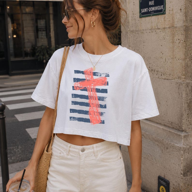 May include: A white short-sleeved crop top featuring a graphic print of a bright coral-colored cross layered over horizontal navy blue stripes. The shirt has a crew neckline and a boxy fit, constructed from a plain white fabric material.