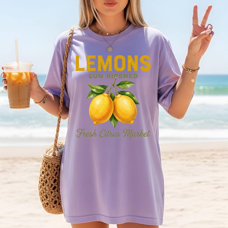 Op de afbeelding: Een lichtpaars t-shirt met korte mouwen en een afbeelding van twee gele citroenen met groene bladeren. De tekst LEMONS SUN RIPENED staat in dikke gele letters boven het fruit en Fresh Citrus Market in groene letters eronder.