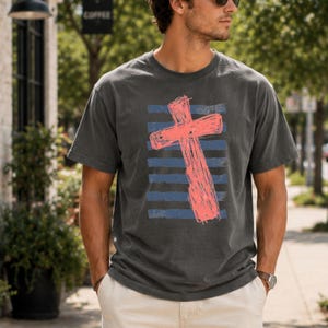 May include: A charcoal gray short-sleeve t-shirt featuring a large, textured coral-colored cross graphic centered over a series of horizontal navy blue stripes. The design has a distressed, hand-drawn appearance on the front of the garment.