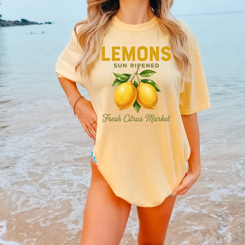 Op de afbeelding: Een lichtgeel T-shirt met korte mouwen en een afbeelding van twee gele citroenen met groene bladeren. De tekst LEMONS SUN RIPENED staat in dikke gele letters en Fresh Citrus Market in een groen cursief lettertype op de voorkant.
