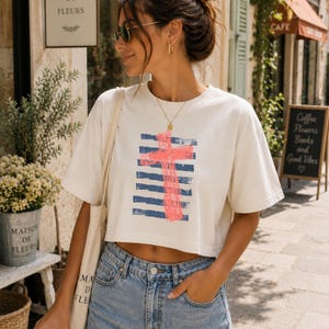 May include: A cream-colored cropped t-shirt featuring a graphic print of a bright pink cross layered over horizontal navy blue stripes. The shirt has short sleeves and a crew neckline, paired with a light-wash denim bottom and a canvas tote bag.