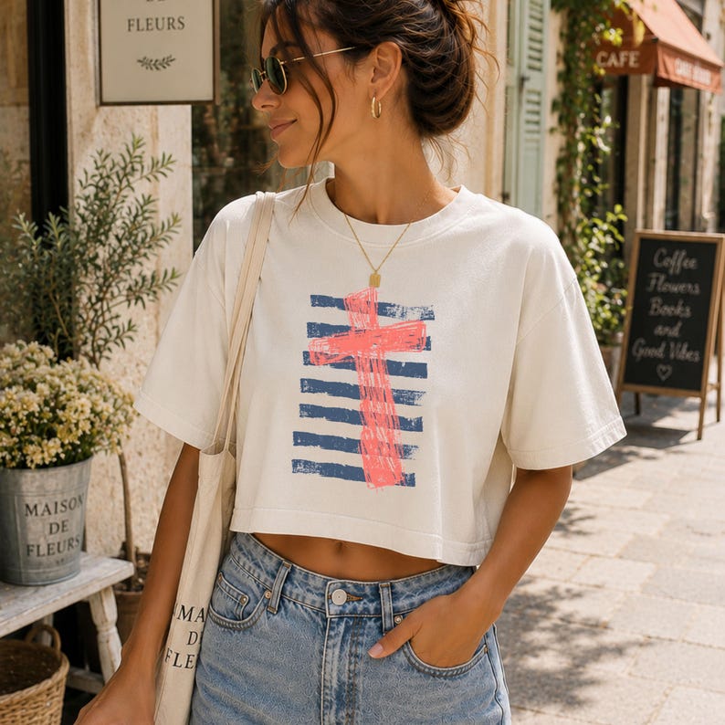May include: A cream-colored cropped t-shirt featuring a graphic print of a bright pink cross layered over horizontal navy blue stripes. The shirt has short sleeves and a crew neckline, paired with a light-wash denim bottom and a canvas tote bag.