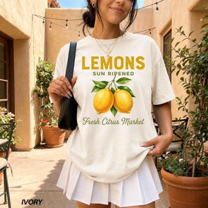 Op de afbeelding: Een ivoorkleurig T-shirt met korte mouwen en een afbeelding van twee gele citroenen met groene bladeren. De tekst luidt LEMONS SUN RIPENED en Fresh Citrus Market in gele en groene letters. Het T-shirt wordt gedragen met een witte plooirok.