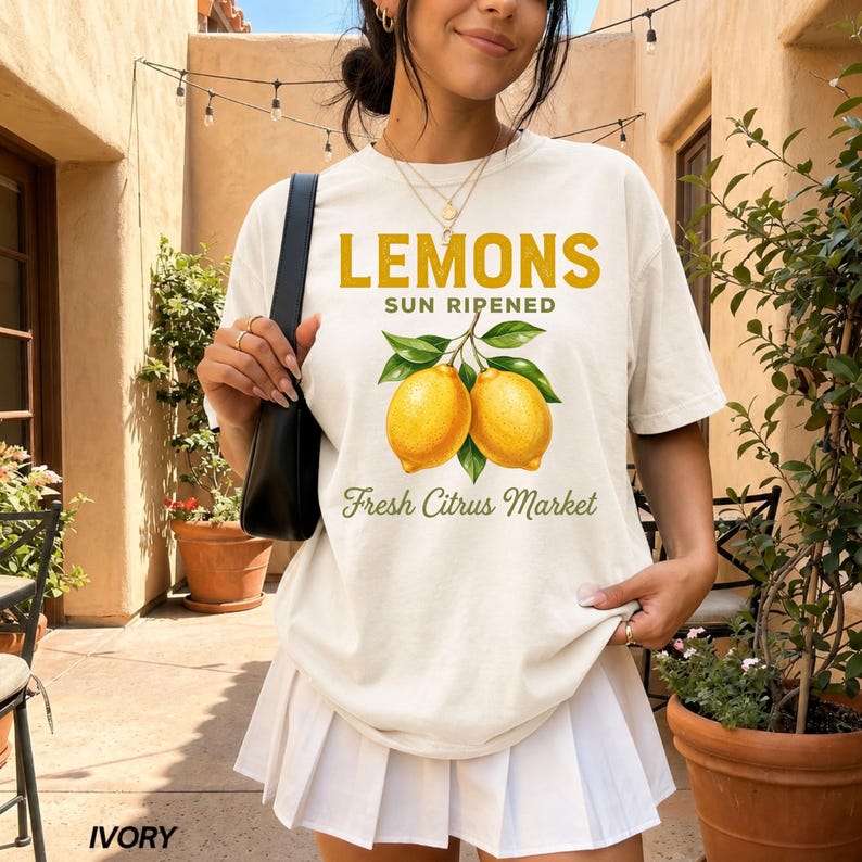 Op de afbeelding: Een ivoorkleurig T-shirt met korte mouwen en een afbeelding van twee gele citroenen met groene bladeren. De tekst luidt LEMONS SUN RIPENED en Fresh Citrus Market in gele en groene letters. Het T-shirt wordt gedragen met een witte plooirok.