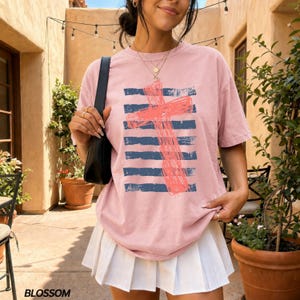 May include: A dusty pink short-sleeve t-shirt featuring a graphic of a coral-colored cross layered over horizontal navy blue stripes. The shirt has a crew neckline and a relaxed fit, paired with a white pleated mini skirt and a black shoulder bag.
