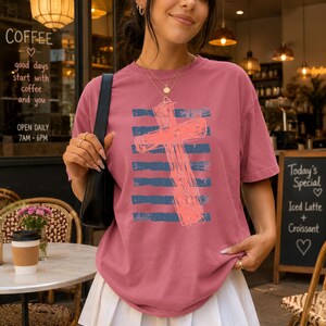 May include: A dusty rose short-sleeve t-shirt featuring a graphic of a coral-colored cross layered over horizontal navy blue distressed stripes. The shirt has a crew neckline and a relaxed fit, worn with a black shoulder bag and layered necklaces.
