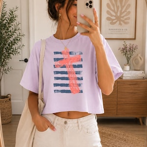May include: A light purple short-sleeved t-shirt featuring a graphic print of a coral-colored cross layered over horizontal navy blue stripes. The shirt has a crew neckline and a relaxed, cropped fit, worn with light-colored denim jeans.