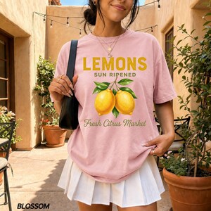 Op de afbeelding: Een lichtroze T-shirt met korte mouwen en een afbeelding van twee gele citroenen met groene bladeren. De tekst LEMONS SUN RIPENED en Fresh Citrus Market staat in een combinatie van dikke gele letters en groen cursief schrift op de voorzijde.