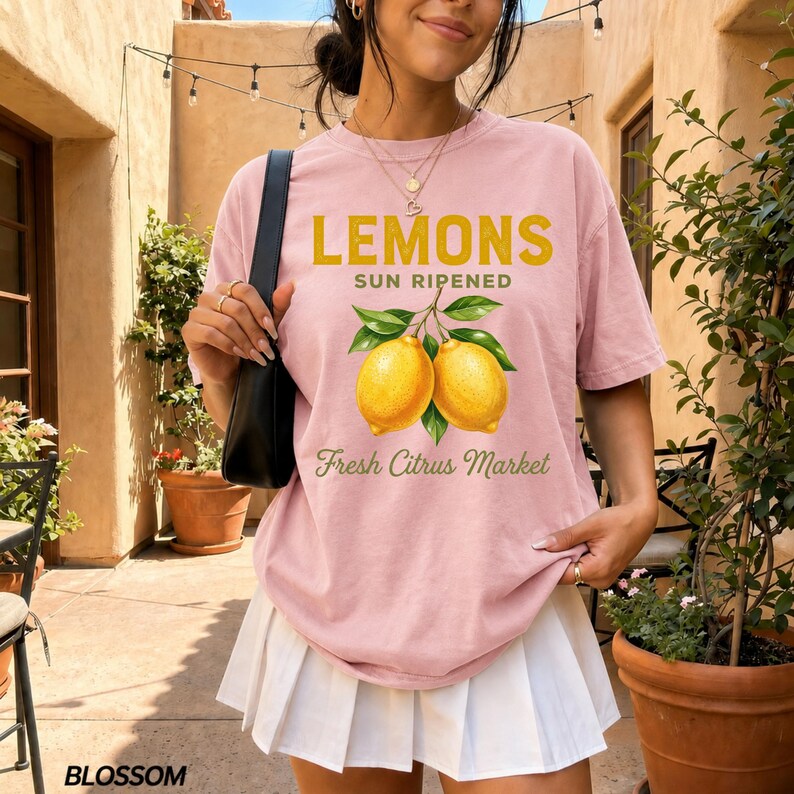 Op de afbeelding: Een lichtroze T-shirt met korte mouwen en een afbeelding van twee gele citroenen met groene bladeren. De tekst LEMONS SUN RIPENED en Fresh Citrus Market staat in een combinatie van dikke gele letters en groen cursief schrift op de voorzijde.