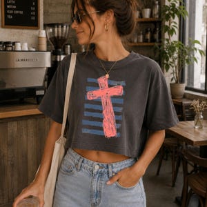 May include: A charcoal gray cropped t-shirt featuring a graphic print of a bright pink cross layered over horizontal blue stripes. The shirt has short sleeves and a crew neckline, paired with light blue denim jeans and a white canvas shoulder bag.