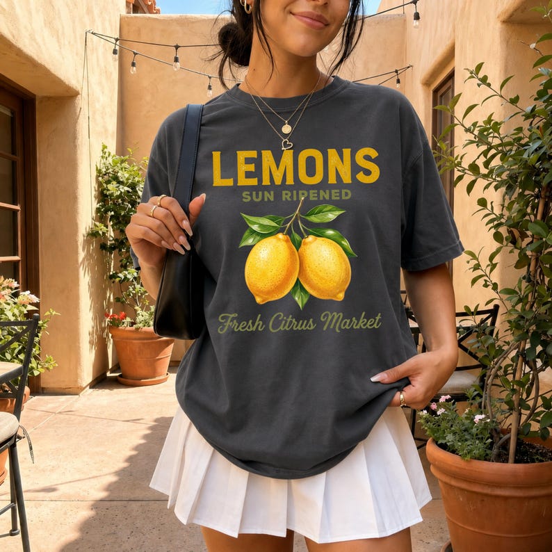 Op de afbeelding: Een antracietgrijs T-shirt met korte mouwen en een afbeelding van twee gele citroenen met groene bladeren. De tekst luidt LEMONS SUN RIPENED en Fresh Citrus Market in gele en groene letters. Het T-shirt wordt gedragen met een witte plooirok.
