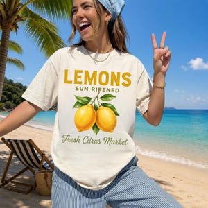 Op de afbeelding: Een cr&egrave;mekleurig T-shirt met korte mouwen en een afbeelding van twee gele citroenen met groene bladeren. De tekst LEMONS SUN RIPENED en Fresh Citrus Market staat in een combinatie van dikke gele en groene lettertypes op de voorzijde.